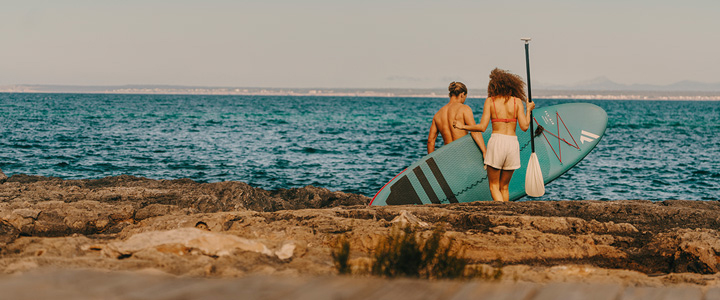 Pärchen läuft am Strand Richtung Meer, Frau trägt Board und Paddel.